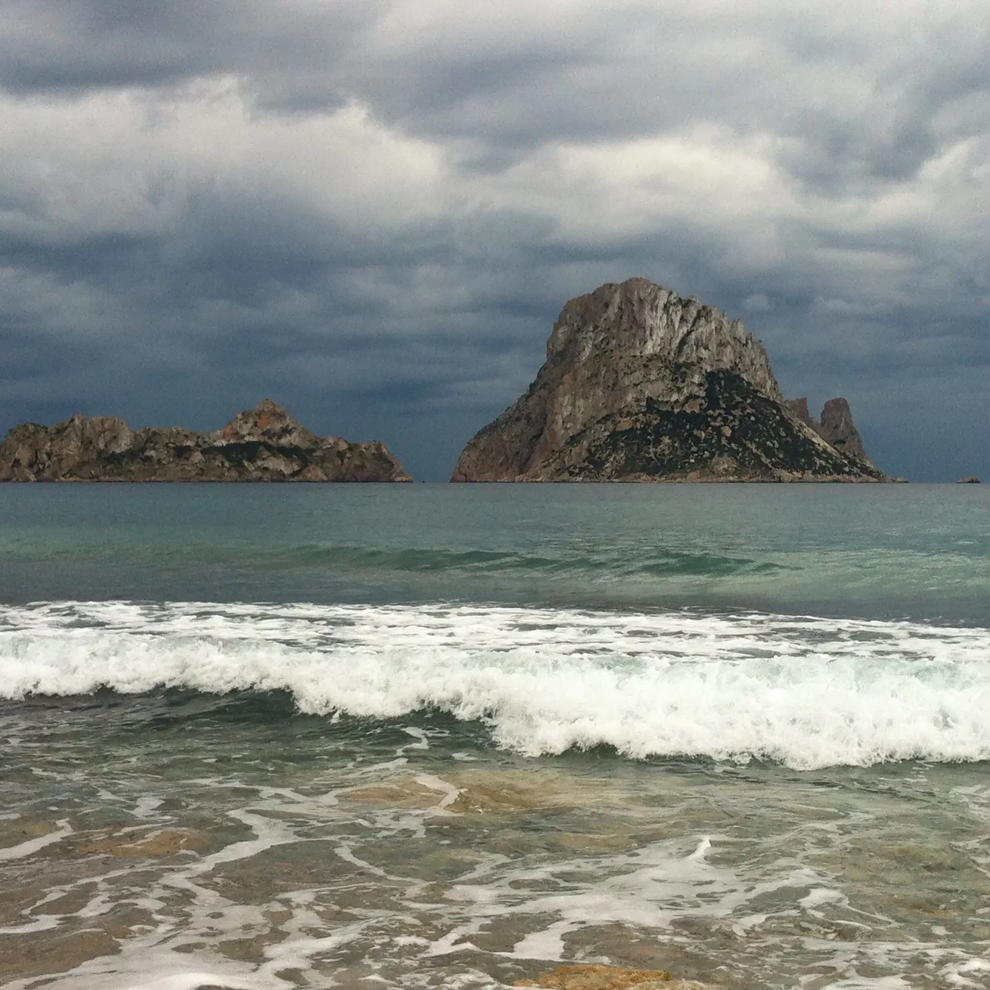 Olas en la orilla de Cala D´Hort con Es Vedrà bajo cielo nublado